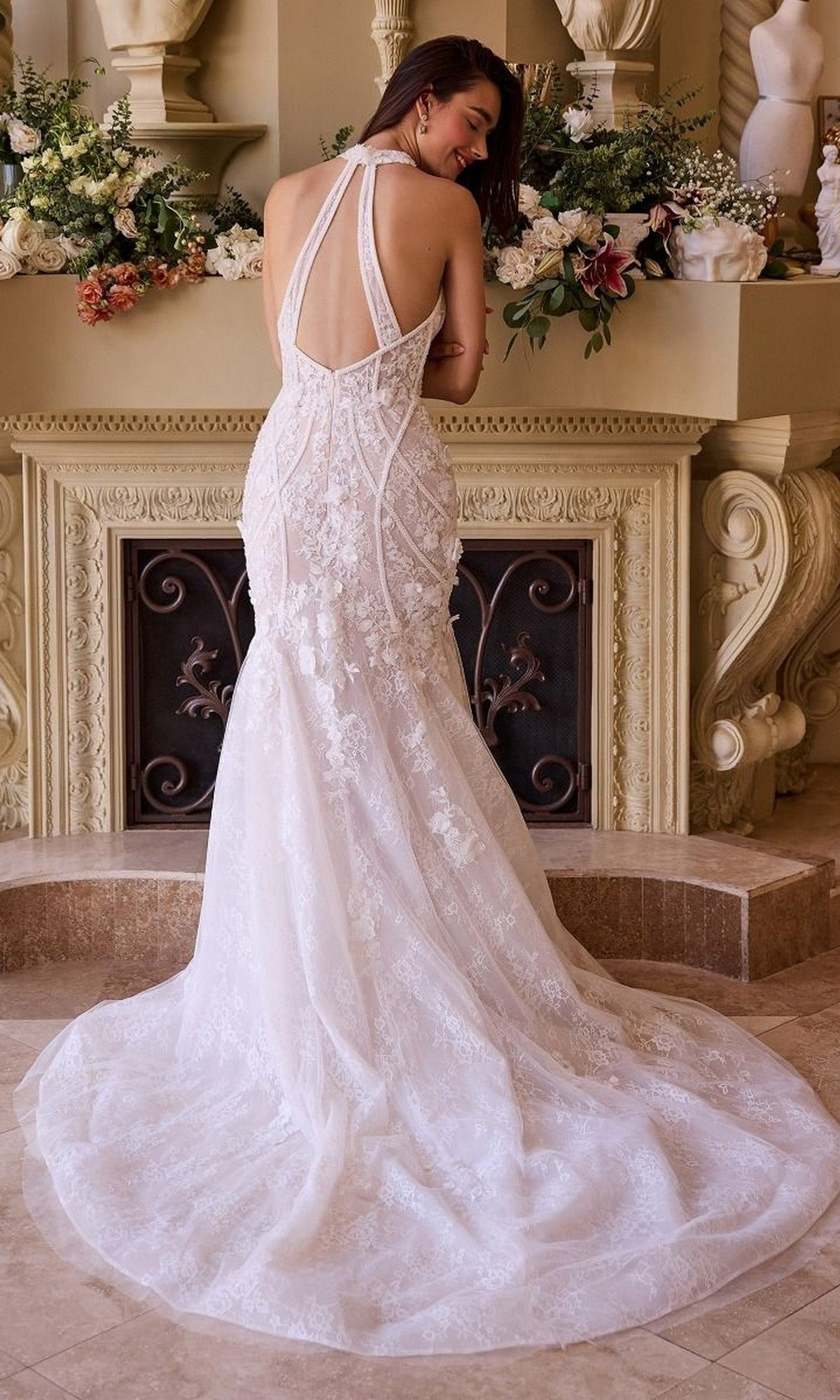 A woman in the Andrea and Leo WL019 long lace, backless mermaid wedding dress with a long train stands before an ornate flower-decorated fireplace.